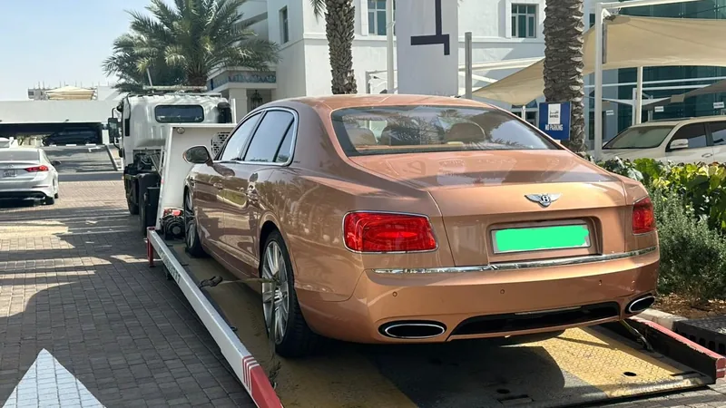 Bronze Bentley Flying Spur sedan being unloaded from recovery flatbed at Dubai luxury hotel valet with palm trees