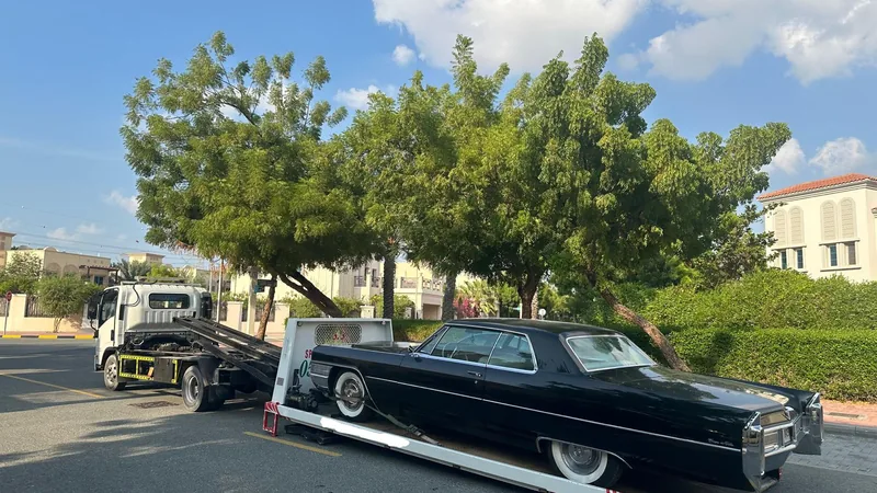 Black classic Cadillac Coupe DeVille loaded on red-and-white recovery flatbed truck on a Dubai villa community street with green trees