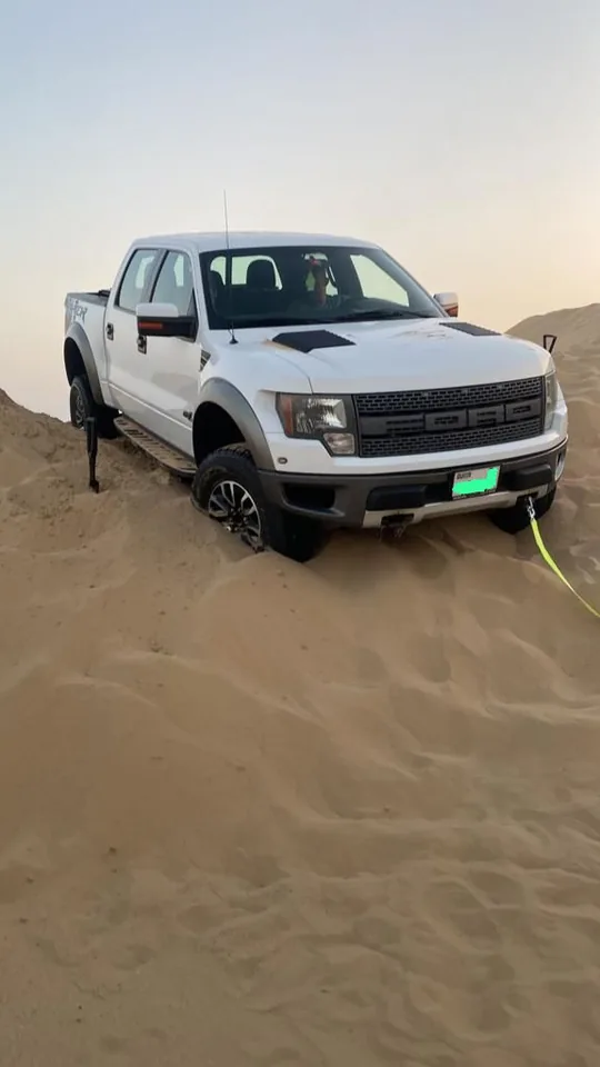 White Ford F-150 Raptor pickup stuck nose-down in soft sand dune in Dubai desert with bright green recovery strap attached